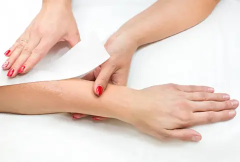 Deux mains avec du vernis rouge retirant une bande d'épilation sur un avant-bras